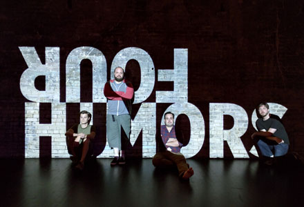 Image of the Four Humors logo projected on a theater wall. The performers are positioned within the letters of the logo.