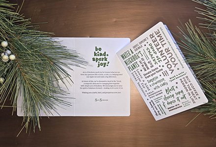 Holiday card on a table with greenery. Headline reads: Be kind. Spark joy!